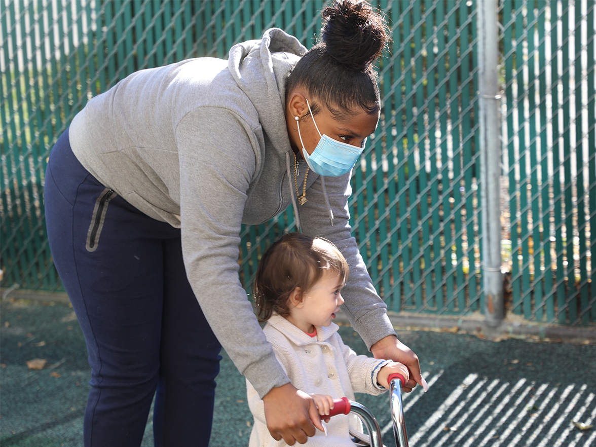 teacher with mask on helping toddler ride a bike outside