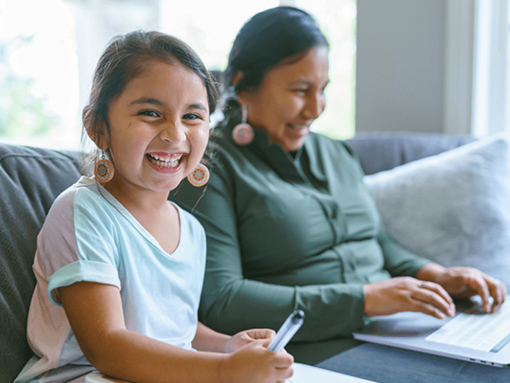 A mother working from home on the couch while her daughter does homework beside her